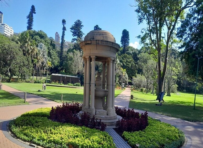 A classical columned structure in a park with a pathway, greenery, and buildings in the background. 5