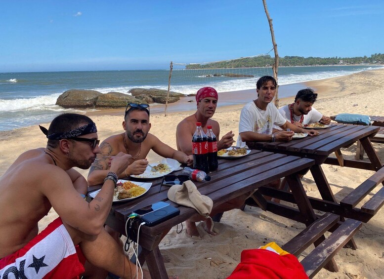 A group of people dining at a wooden table on a sandy beach with ocean waves in the background. 5