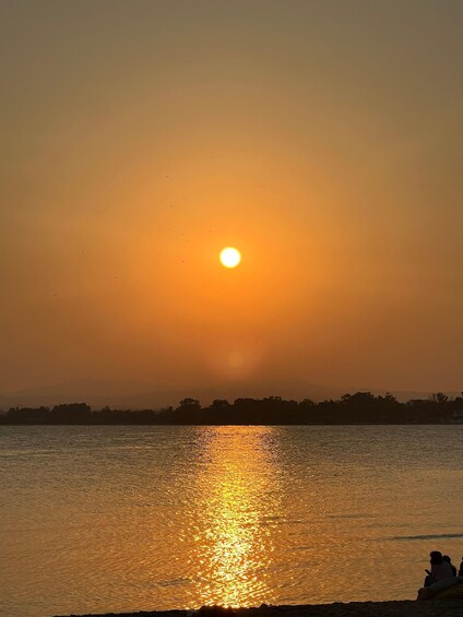 A sunset over a calm body of water with a silhouette of a distant shoreline and a few people sitting by the water's edge. 1