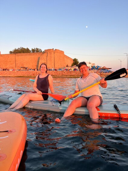 Two people sitting on paddleboards in calm water with a stone wall and a clear sky in the background. 3