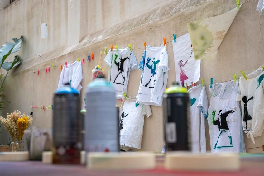 A wall with clothes hanging on a line, some with drawings, and two large containers on the ground. 2