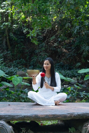 A person sitting on a stone bench in a lush garden, holding a bowl and a red flower. 3