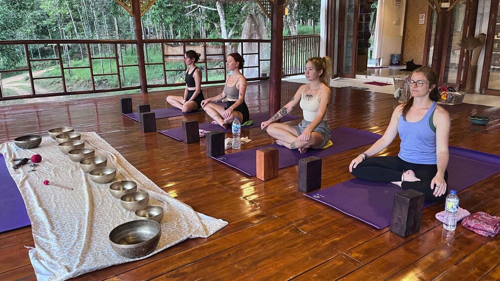 Four people meditating on yoga mats in a room with wooden floors and large windows. 1