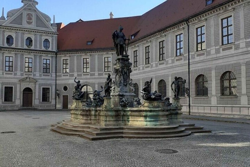 A historical building with classical architecture, a central fountain with statues, and a cobblestone courtyard. 1