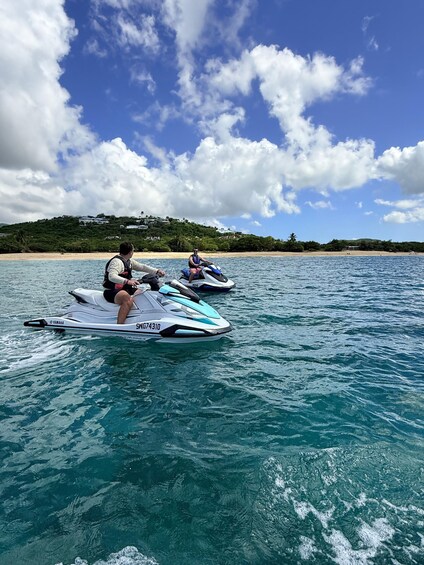 Two people jet-skiing on a clear day with a beach and hills in the background. 2