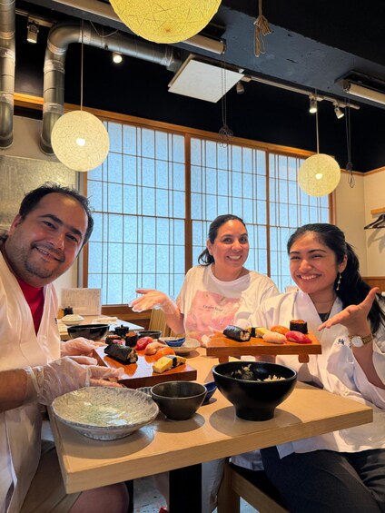 Three people at a table with various sushi dishes and bowls, in a room with large windows and hanging lights. 3