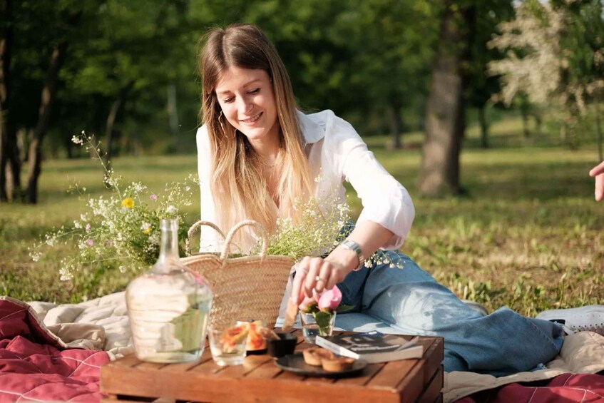 A person sitting on a blanket in a park with a picnic setup including a basket, a bottle, and food. 1