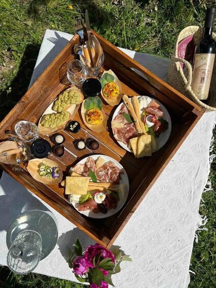 A picnic setup with a wooden tray containing various cheeses, meats, and small dishes, set on a white cloth outdoors. 2