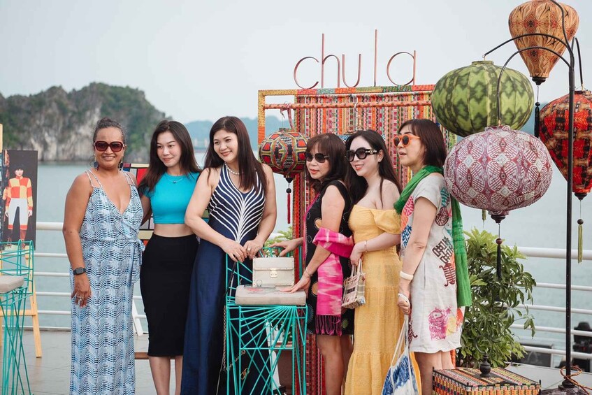A group of people standing in front of a colorful 'chula' sign with decorative lanterns and a scenic waterfront in the background. 2
