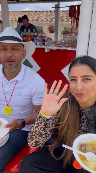 Two people seated at an outdoor table with food, a red and white tablecloth, and a tiled roof in the background. 3