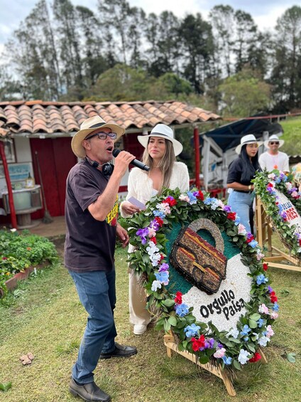 A man speaking into a microphone, a woman holding a floral wreath, and another person in the background. 5