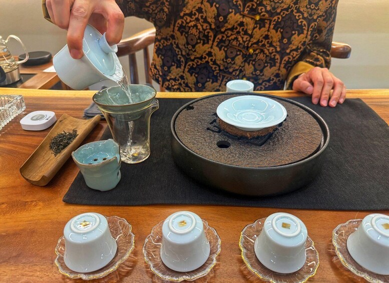 A person pouring tea into a glass, with a teapot and tea leaves on a wooden tray, and several teacups on the table. 3