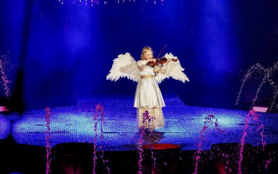 A person playing a violin on a stage with a blue background and white feathers. 2