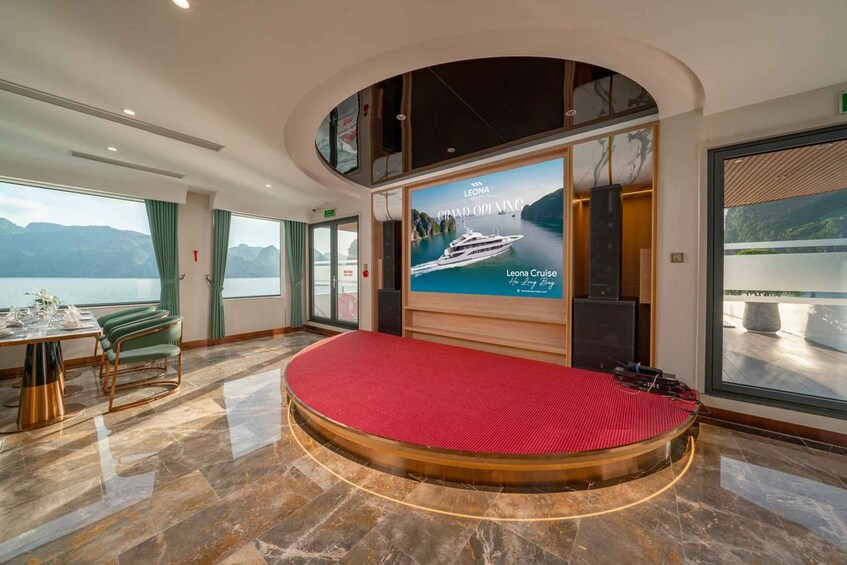A cruise ship interior with a large screen displaying a yacht, a round red carpet, a dining area with green chairs, and a view of the sea through large windows. 2