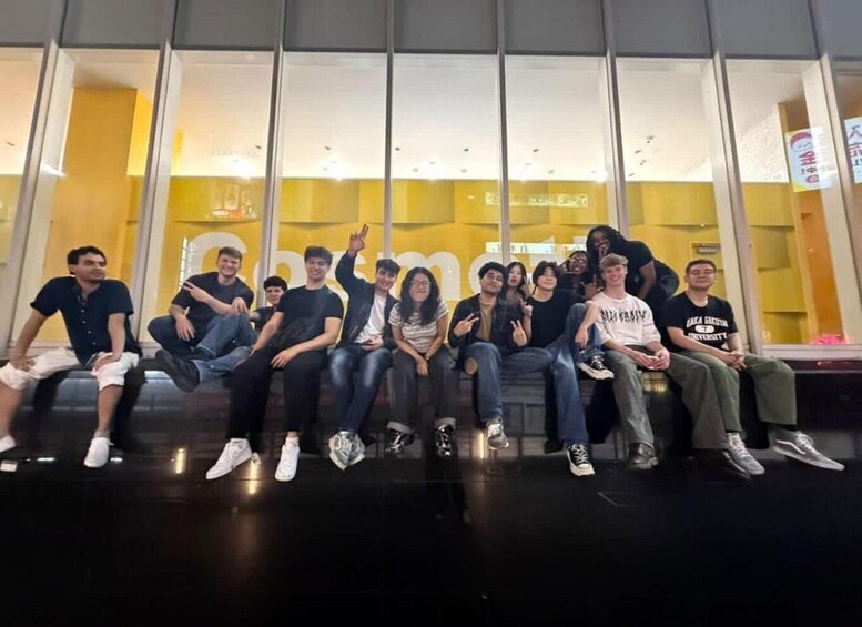 A group of people sitting on a bench in an indoor setting with large windows and a yellow wall in the background. 3