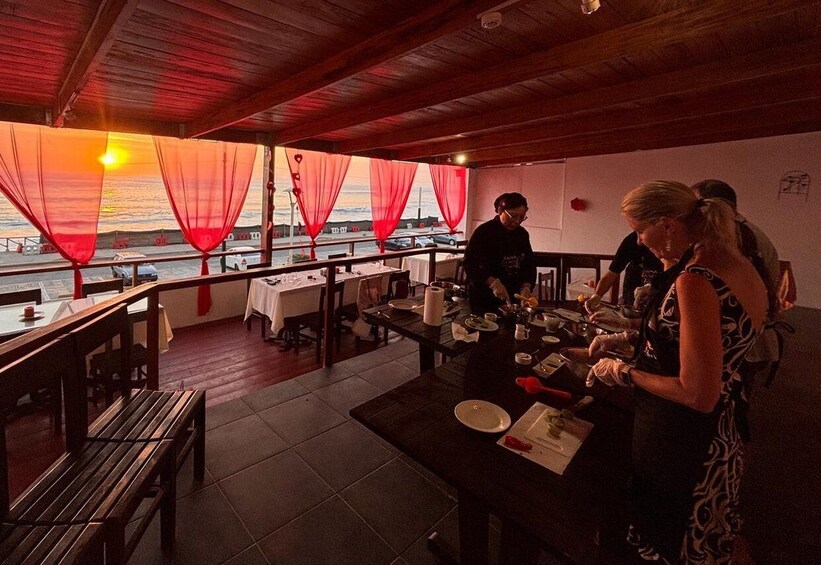 Un restaurant en plein air avec des tables et des bancs en bois, des rideaux rouges et une vue sur le coucher de soleil sur l’eau. 1