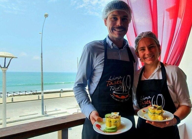 Deux personnes portant des tabliers arborant le logo « Lima Tasty Tours », tenant des assiettes de nourriture, avec une plage et l’océan en arrière-plan. 5