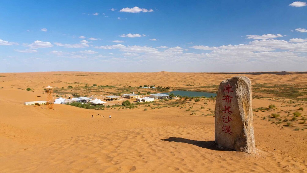 A desert landscape with a large rock inscribed with text, a small settlement with white buildings, and a body of water in the distance. 2
