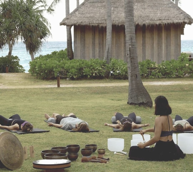 A group of people meditating outdoors with thatched-roof huts and palm trees in the background. 1