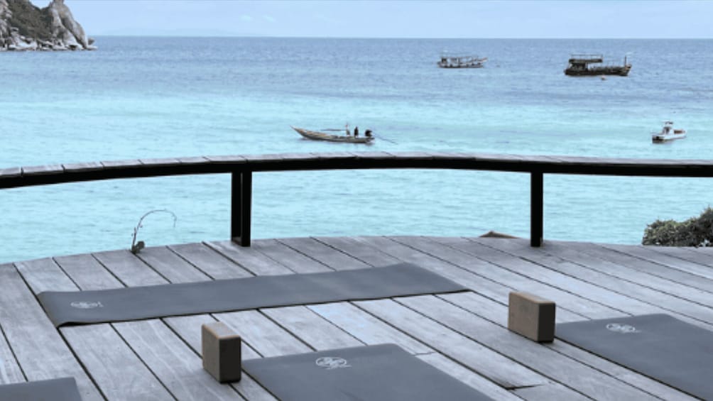 A wooden deck with yoga mats and a railing overlooking turquoise waters with boats. 2