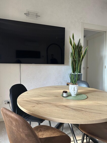 A round wooden table with a white vase and a plant, a TV mounted on the wall, and a door in the background. 3