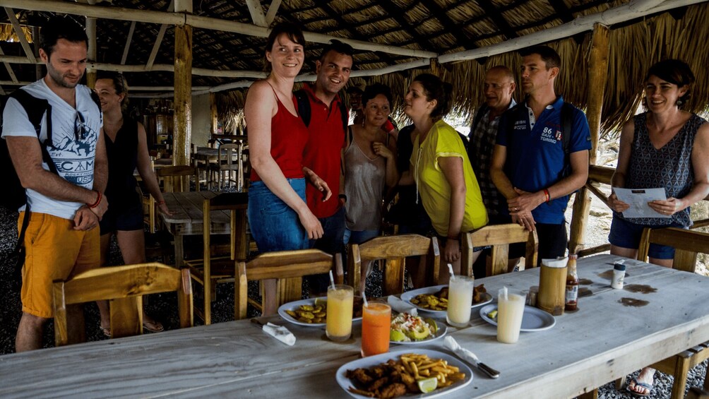 A group of people gathered around a table with food and drinks, under a thatched roof structure. 2