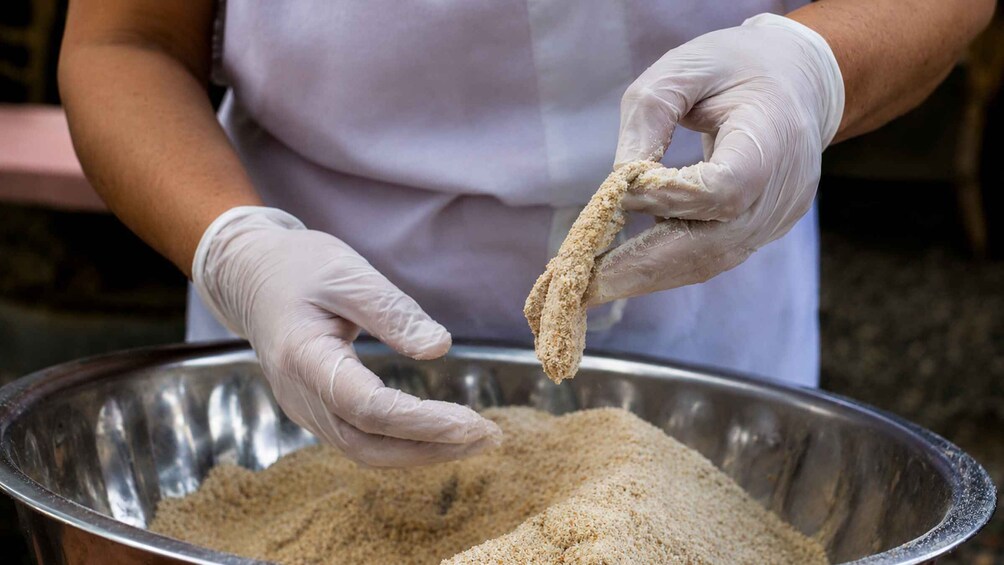A person wearing gloves holding a breaded item over a large metal bowl filled with a granular substance. 4