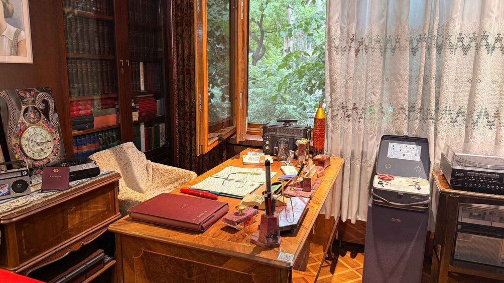 A vintage study room with a wooden desk, bookshelves, a radio, a typewriter, and a window with lace curtains. 4