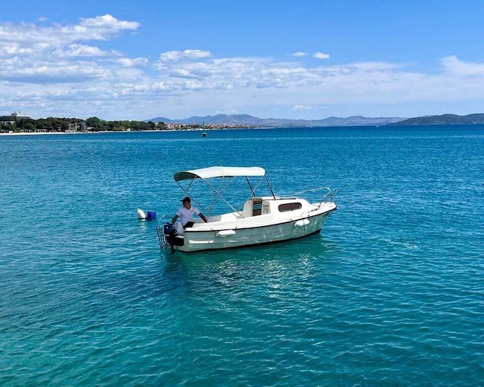 A small boat with a canopy on calm turquoise waters, a person on board, and a distant shoreline with buildings and mountains. 1