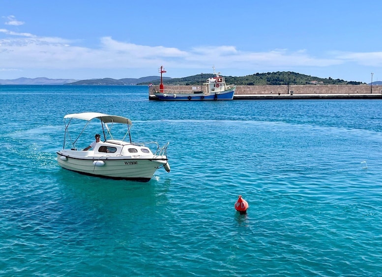 A small white boat with a canopy on a calm sea, a red buoy, and a larger blue boat in the background near a stone pier. 5