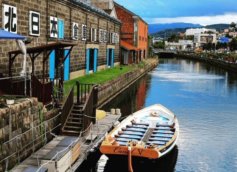 A canal with a boat docked at a wooden pier, a stone building with blue doors, and a bridge in the background. 4
