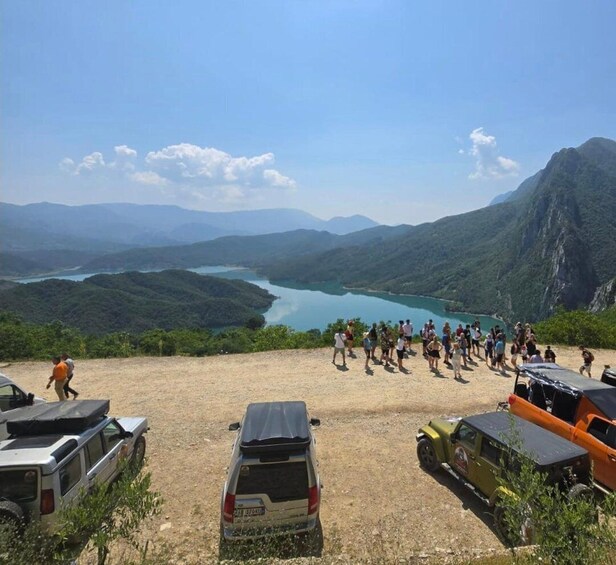 A group of people walking on a dirt path with vehicles parked nearby, overlooking a lake and mountains. 1
