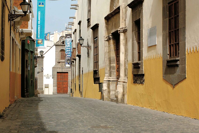 A narrow cobblestone street flanked by historic buildings with yellow and white facades, a church with a bell tower, and banners hanging from the buildings. 2