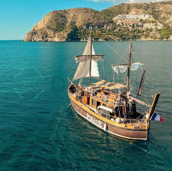 A wooden sailing boat named 'Pirate' on calm waters with a hilly coastline in the background. 1