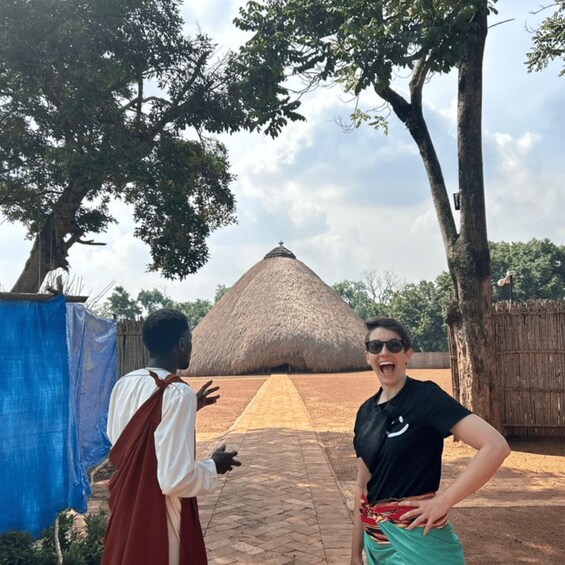Two people standing in front of a thatched-roof structure with a paved pathway leading to it. 1