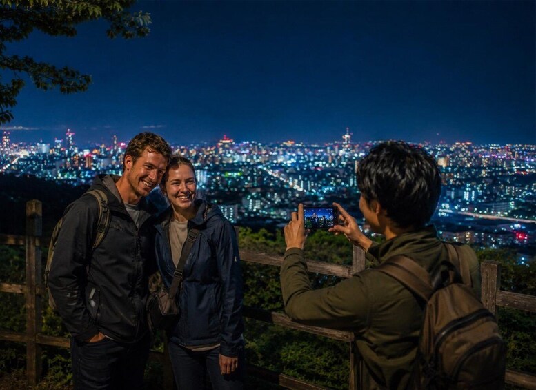 Three people standing by a railing with a cityscape at night in the background. 3