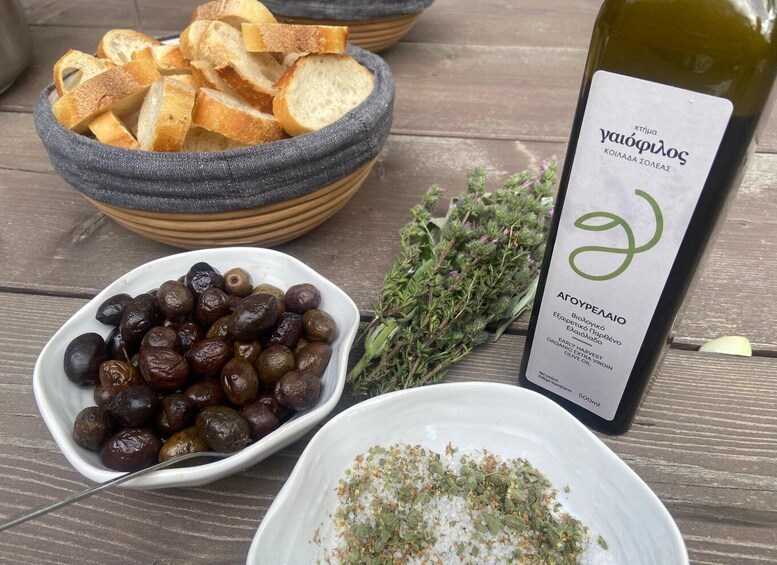 A bottle of Aroyomenai wine, a bowl of olives, a plate of bread, and some herbs on a wooden table. 5
