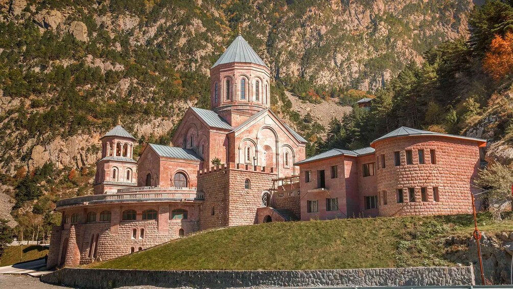 A large, multi-story building with a red brick facade, multiple towers, and a steep hillside backdrop. 1