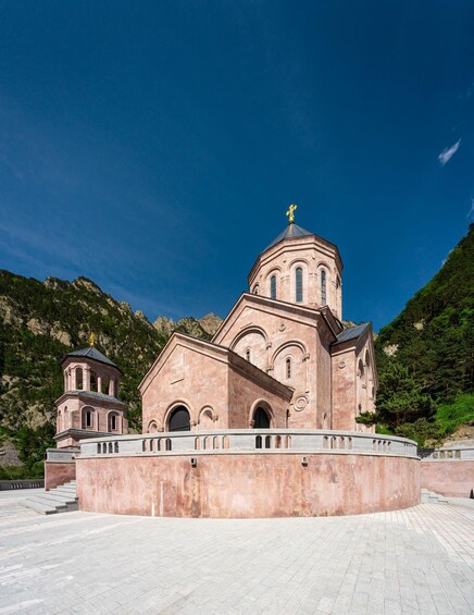A large, pinkish-red church with a golden dome and cross, surrounded by a paved courtyard and lush green hills. 2