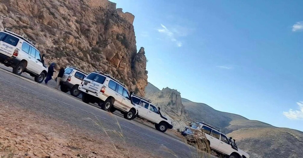 A line of white SUVs parked on a road with a rocky cliff in the background. 1