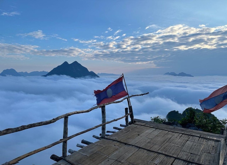A wooden viewing platform with a flag, overlooking a sea of clouds and distant mountains under a partly cloudy sky. 1
