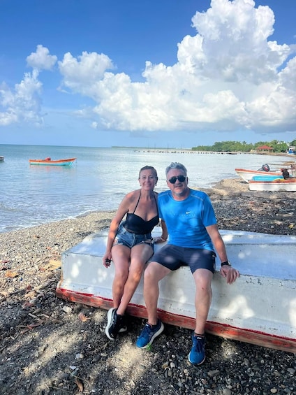 Two people sitting on a concrete bench by a beach with boats and a pier in the background. 2