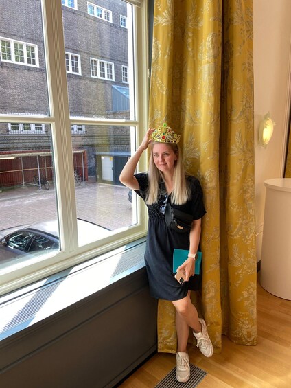 A person wearing a crown, sitting by a window with yellow curtains, holding a blue book, and wearing a black dress with a white belt. 2