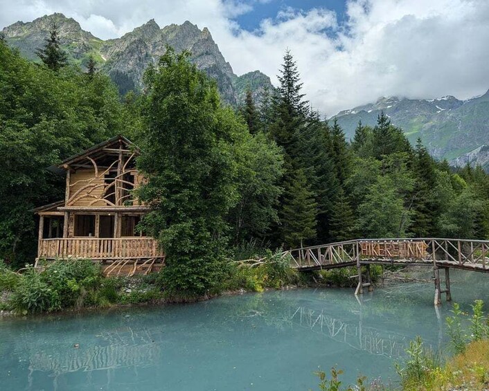 A wooden cabin by a turquoise lake with a wooden bridge, surrounded by dense forest and mountains. 1