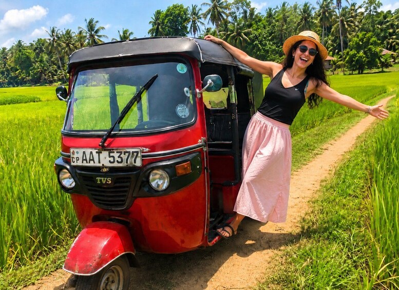 A red auto-rickshaw parked on a dirt path with a woman standing beside it, wearing a hat and a pink skirt, in a lush green field with palm trees in the background. 1