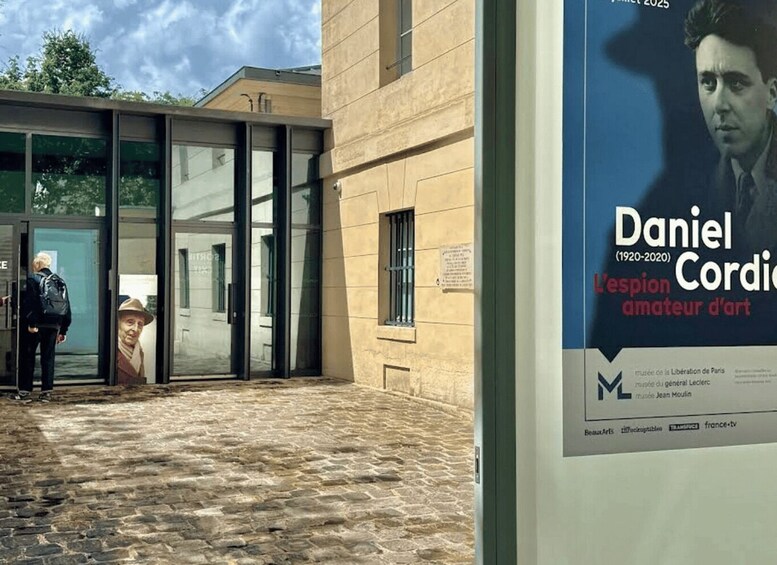 A building entrance with glass doors, a cobblestone path, and a poster advertising an art exhibition. 3