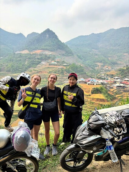 Three people standing by a motorcycle with a scenic mountainous landscape in the background. 4