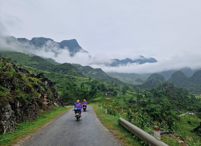 Two motorcyclists on a paved road surrounded by lush green hills and mountains. 1