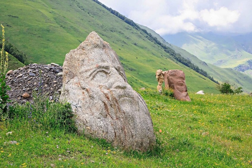A large rock with a carved face, a smaller rock, and a grassy hillside with a mountain in the background. 4