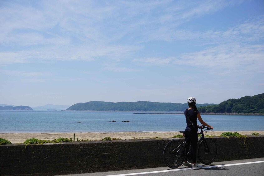 A person on a bicycle by a coastal road with a view of the sea and distant hills. 1